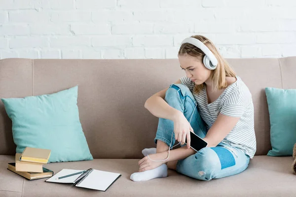 Triste adolescente chica sentado en sofá y escuchar música con auriculares - foto de stock