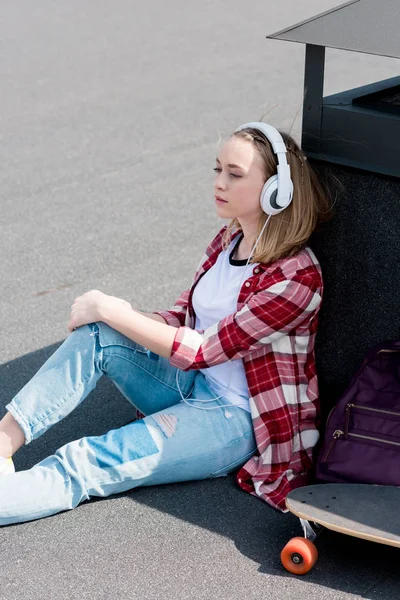 Schöne Teenie-Mädchen mit Skateboard sitzt auf dem Dach und hört Musik mit Kopfhörern — Stockfoto