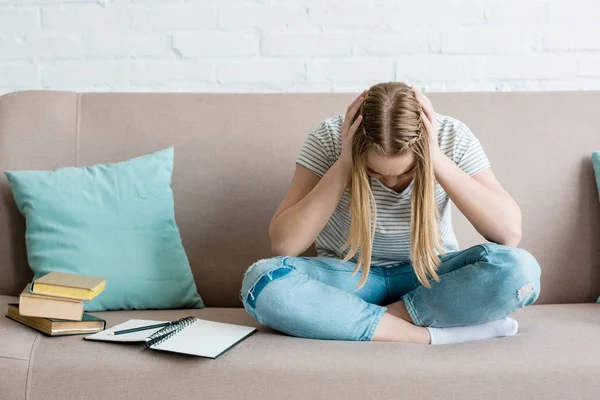 Depressed teen girl sitting on couch with books and holding head — Stock Photo