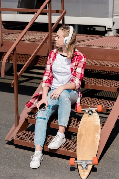 Beautiful teen girl in red plaid shirt and headphones sitting on stairs with skateboard — Stock Photo