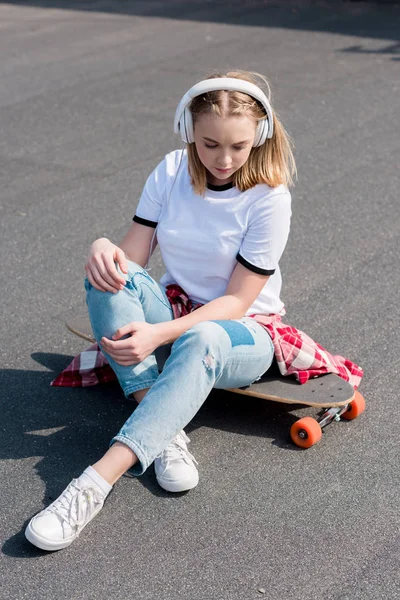 Stylish teen girl listening music in headphones while sitting on skateboard — Stock Photo