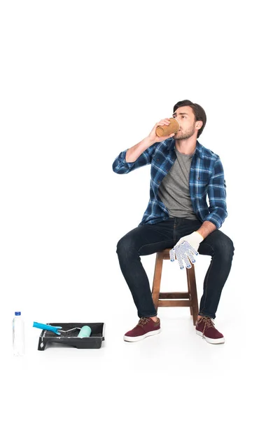 Чоловік відпочиває на стільці і п'є каву біля роликового лотка і фарбує ролик ізольовано на білому тлі — стокове фото