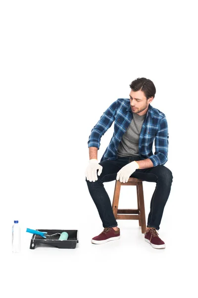 Hombre en guantes de protección descansando en silla cerca de bandeja de rodillo y rodillo de pintura aislado sobre fondo blanco - foto de stock