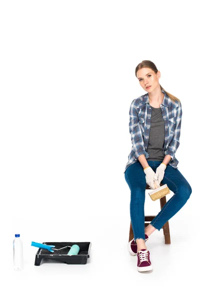 Mujer joven sentada en silla con pincel de pintura cerca de botella, bandeja de rodillo y rodillo de pintura aislados sobre fondo blanco - foto de stock