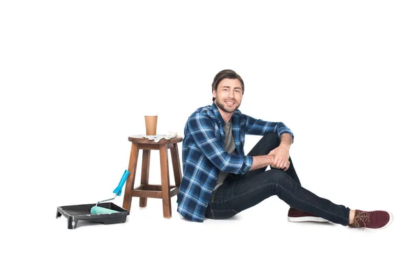 Homme assis près de la chaise avec tasse de café et plateau à rouleaux avec rouleau de peinture isolé sur fond blanc — Photo de stock