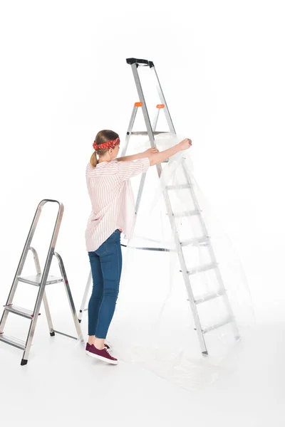 Rückansicht einer Frau mit Stirnband, die von einer Leiter die Polyethylenabdeckung abnimmt, isoliert auf weißem Hintergrund — Stockfoto