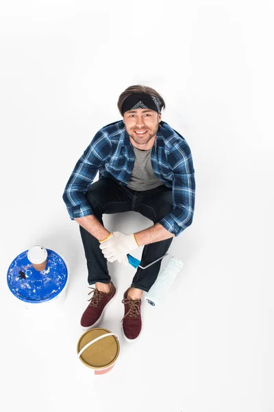 Vista de ángulo alto del hombre en diadema y guantes protectores sosteniendo rodillo de pintura y sentado en el suelo con latas de pintura y taza de papel de café aislado sobre fondo blanco - foto de stock