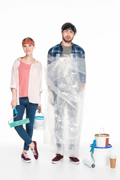 Smiling woman with paint tin and paint roller standing with boyfriend in polyethylene cover near painting tools and coffee cup isolated on white background — Stock Photo