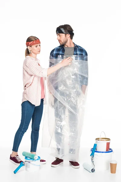 Jeune femme enlevant la couverture de polyéthylène de petit ami et outils de peinture avec tasse de café sur le sol isolé sur fond blanc — Photo de stock