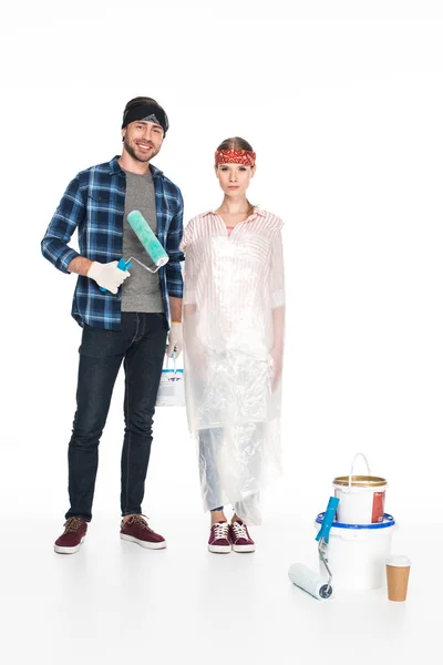 Jeune homme souriant avec rouleau de peinture debout près de petite amie en polyéthylène couverture et outils de peinture avec tasse en papier de café sur le sol isolé sur fond blanc — Photo de stock