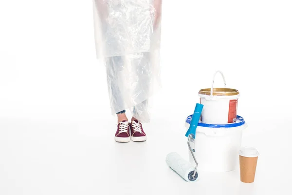 Image recadrée de la femme dans une couverture en polyéthylène debout près de boîtes de peinture, rouleau de peinture et tasse de papier de café isolé sur fond blanc — Photo de stock