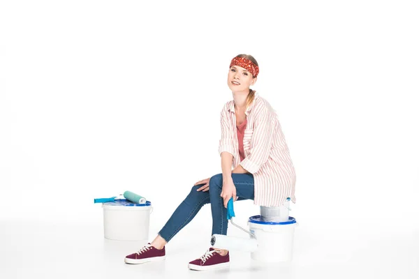 Woman in headband sitting with paint roller on paint tins isolated on white background — Stock Photo