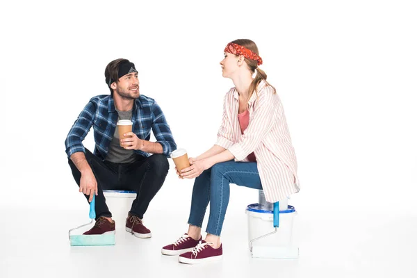 Усміхнена молода пара сидить на пляшках з фарбою і п'є каву з паперових стаканчиків ізольовано на білому тлі — стокове фото