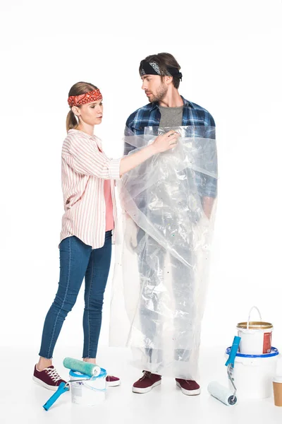 Young woman taking off polyethylene cover from boyfriend and painting tools with coffee cup on floor isolated on white background — Stock Photo