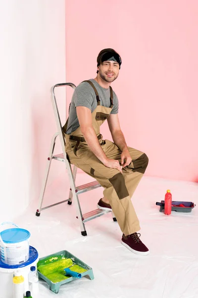 Homem em geral trabalhando sentado na escada perto de bandejas de rolo com rolos de tinta e latas de tinta — Fotografia de Stock
