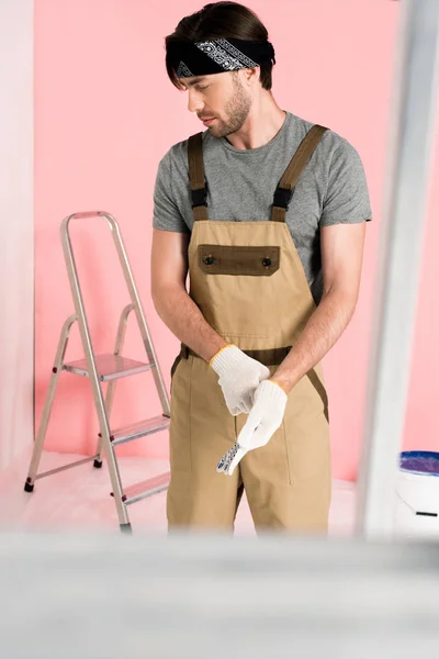 Молодой человек в работе в целом надевая защитные перчатки в комнате с лестницами и краской олова — стоковое фото