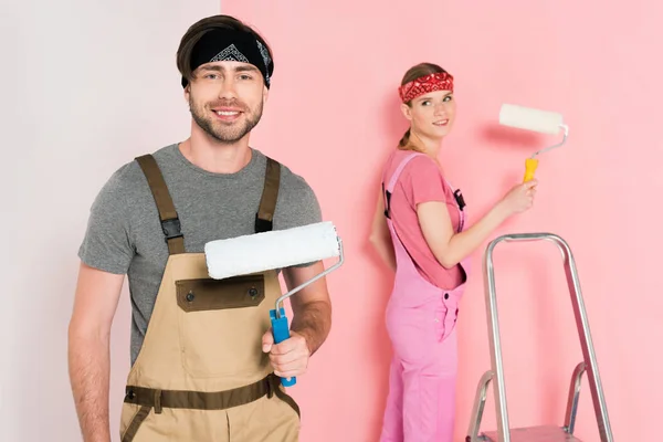 Sonriente hombre con rodillo de pintura y su novia de pie detrás y pared de pintura - foto de stock