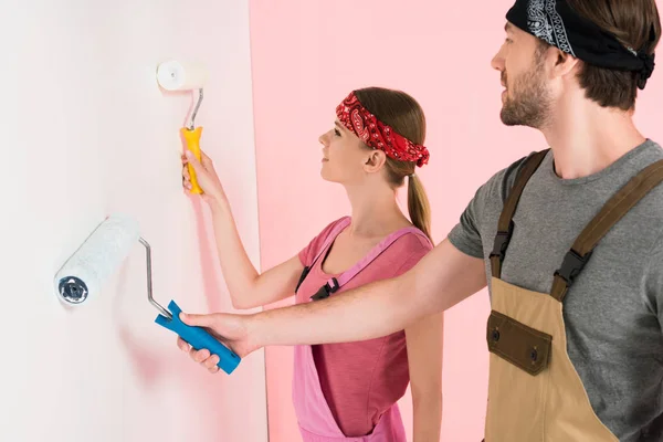 Vue latérale du jeune couple en salopette de travail peinture murale par rouleaux de peinture — Photo de stock