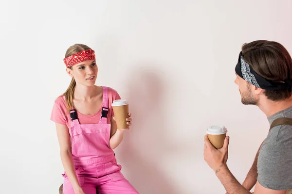 Молодая пара в повязках и рабочем комбинезоне пьет кофе из бумажных стаканчиков — стоковое фото