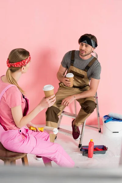 Mujer descansando en silla con taza de papel de café y novio sentado en escalera con taza de café en la habitación con herramientas de pintura - foto de stock