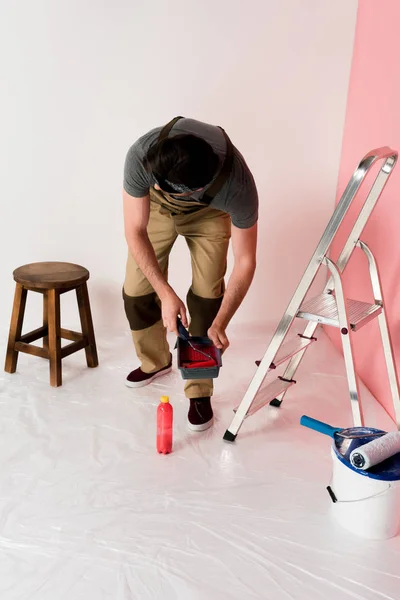 Front view of man in working overall and headband dipping paint roller into roller tray — Stock Photo