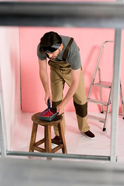 Hombre en general de trabajo y diadema de inmersión rodillo de pintura en bandeja de rodillos en silla - foto de stock