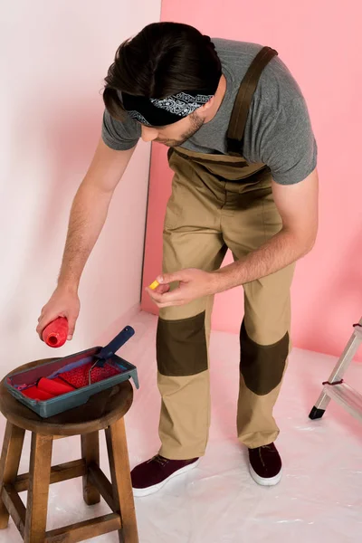 Jeune homme dans le travail en général et serre-tête verser de la peinture de la bouteille dans le plateau à rouleaux sur la chaise — Photo de stock