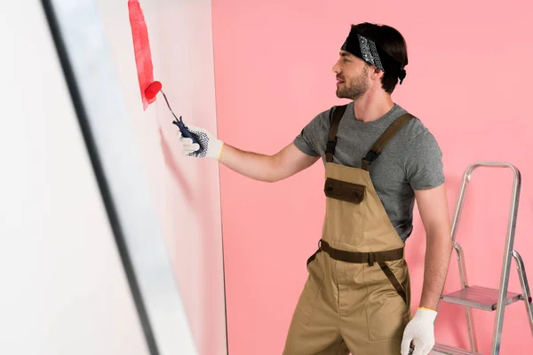 Side view of man in working overall and headband painting wall in red by paint roller near ladder — Stock Photo