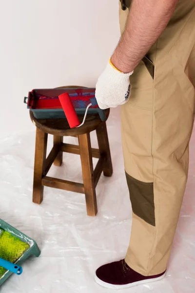 Colpo ritagliato di uomo nel funzionamento complessivo tenuta rullo di vernice e in piedi vicino vassoio rullo sulla sedia — Foto stock