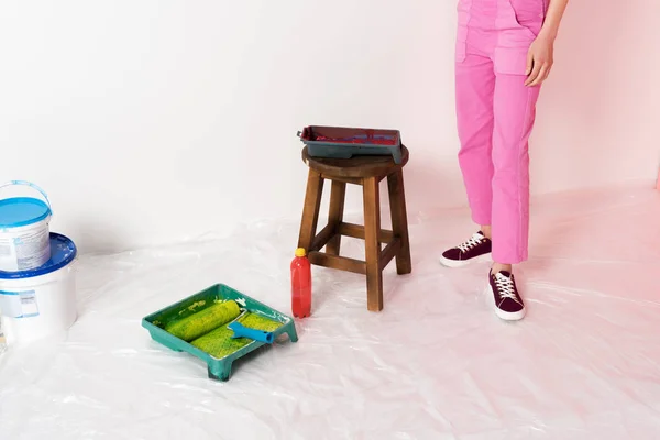 Cropped shot of woman in working overall standing near chair, roller trays, paint tins and bottle — Stock Photo