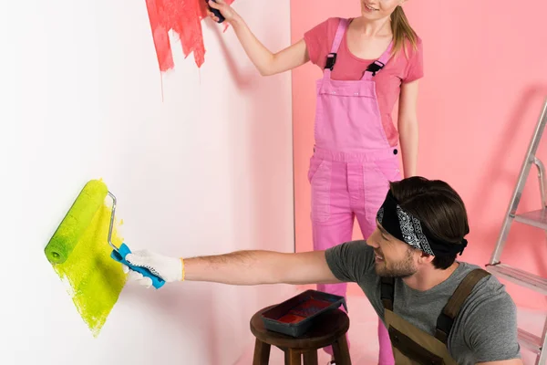 Pareja en overoles de trabajo pintura pared por rodillos de pintura cerca de escalera - foto de stock