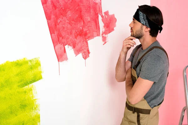 Thoughtful young man in working overall looking at half painted wall and holding hand on chin — Stock Photo