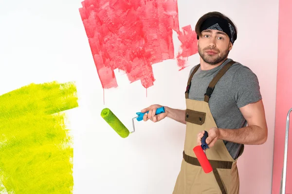 Young man in working overall holding paint rollers and doing shrug gesture in front of painted wall — Stock Photo