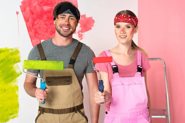 Sonriente pareja joven en overoles de trabajo con caras pintadas sosteniendo rodillos de pintura - foto de stock