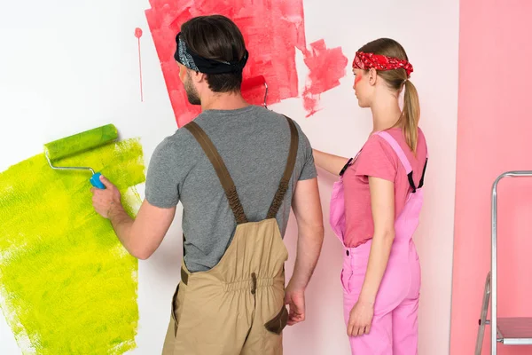 Rückansicht eines jungen Paares in Arbeitskleidung, das die Wand mit Farbwalzen nahe der Leiter bemalt — Stockfoto