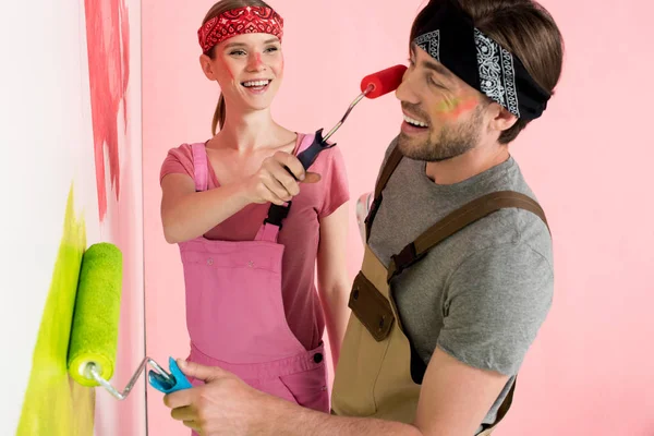 Happy woman in working overall painting face of smiling boyfriend by paint roller while he painting wall — Stock Photo