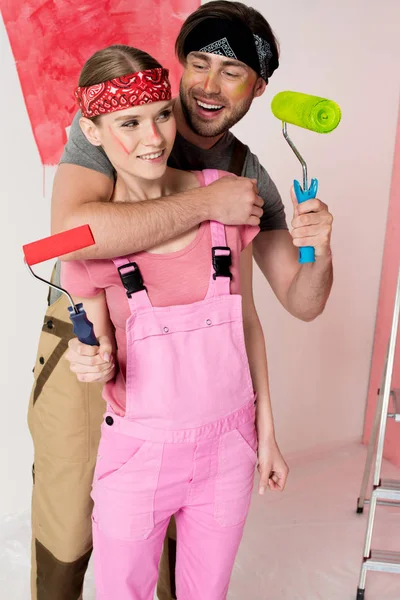 Smiling man with paint roller embracing girlfriend in working overall in front of half painted wall — Stock Photo