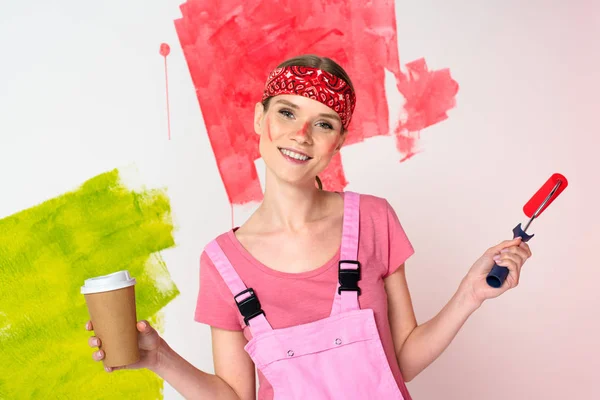 Femme souriante avec visage peint tenant tasse de papier de café et rouleau de peinture — Photo de stock