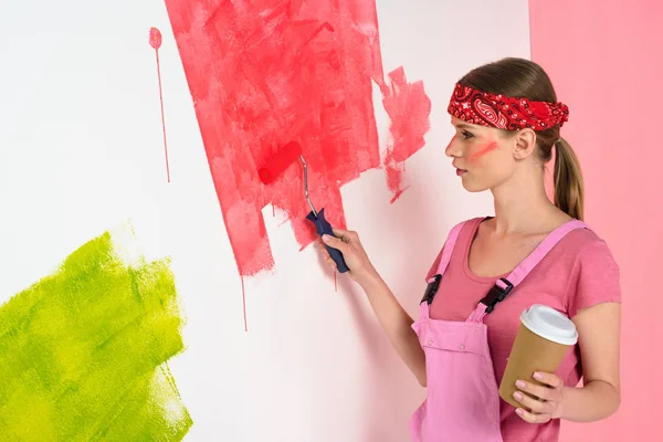 Mujer joven con taza de café pintura pared en rojo por rodillo de pintura - foto de stock