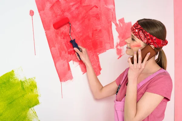 Vista lateral de la mujer sonriente hablando en el teléfono inteligente y pintando la pared en rojo por rodillo de pintura - foto de stock