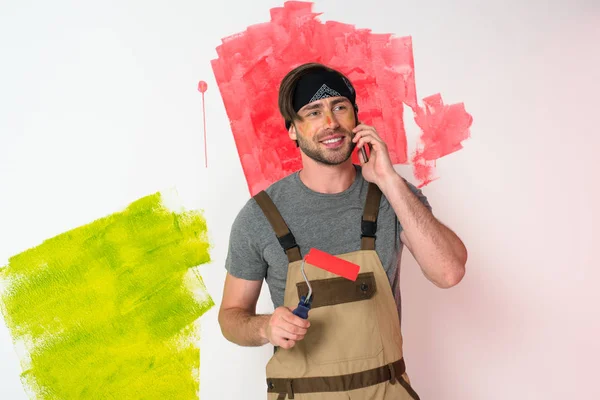 Joven en diadema hablando en el teléfono inteligente y sosteniendo rodillo de pintura delante de la pared pintada - foto de stock