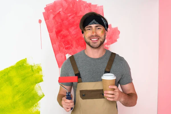 Sonriente hombre con cara pintada sosteniendo rodillo de pintura y taza de papel de café - foto de stock