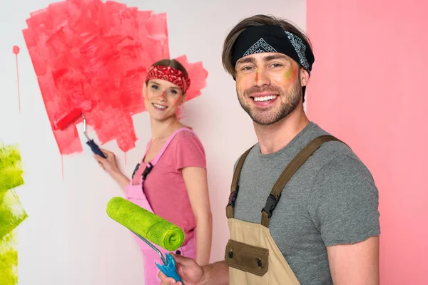 Sonriente pareja joven en overoles de trabajo con caras pintadas sosteniendo rodillos de pintura - foto de stock