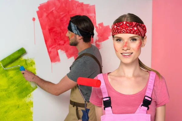 Retrato de jovem mulher com rosto pintado segurando rolo de pintura e seu namorado pintura parede atrás — Fotografia de Stock