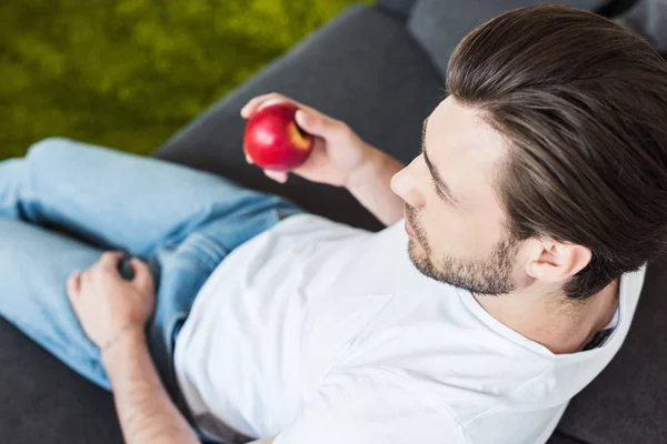 Высокий угол зрения молодого человека, сидящего дома на диване с яблоком в руке — стоковое фото