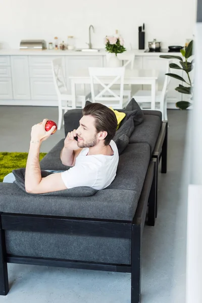 Вид сбоку человека, говорящего по смартфону и сидящего дома на диване с яблоком в руке — стоковое фото