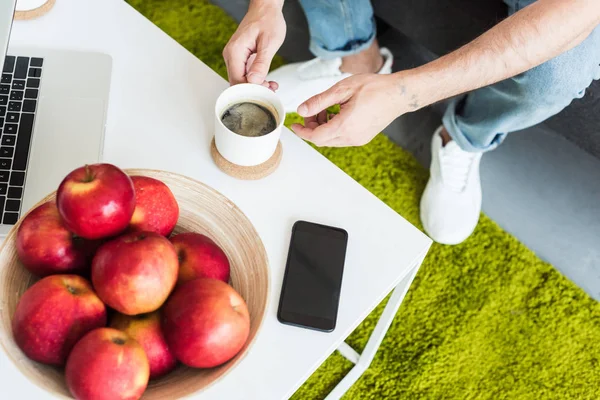 Visão parcial do homem tomando xícara de café da mesa com smartphone, laptop e maçãs em casa — Fotografia de Stock