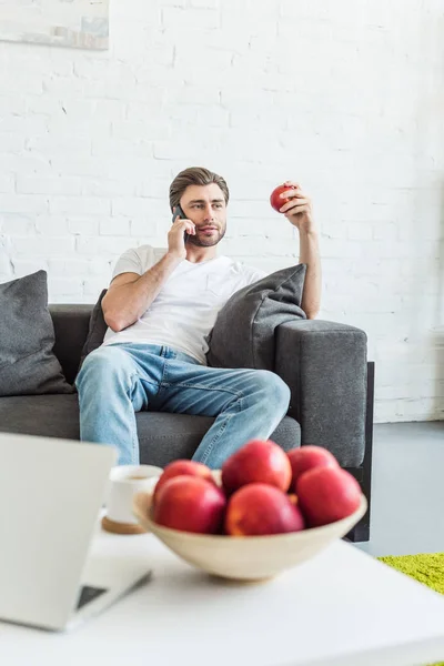 Hombre hablando en el teléfono inteligente y sentado en el sofá con la manzana en la mano en casa - foto de stock