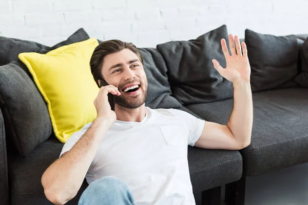 Jovem gesticulando à mão e falando no smartphone perto do sofá em casa — Fotografia de Stock