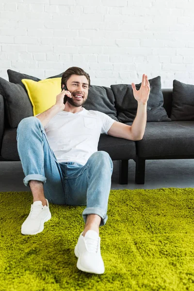 Joven sentado en el suelo y haciendo gestos con la mano mientras habla en el teléfono inteligente en casa - foto de stock